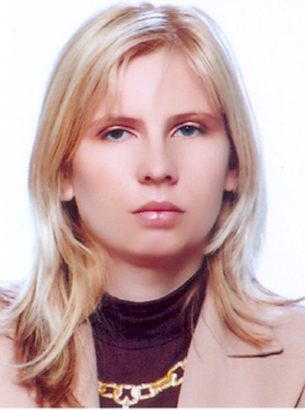 Person with blonde hair, wearing a beige blazer over a black top with a gold chain necklace.